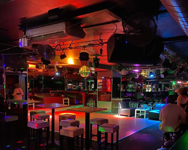 A dimly lit bar features colorful lights and a dance floor surrounded by stools and tables.