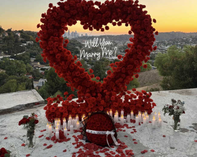 The image shows a large heart-shaped arrangement of red roses surrounded by candles and scattered rose petals, set against a sunset view of a cityscape.