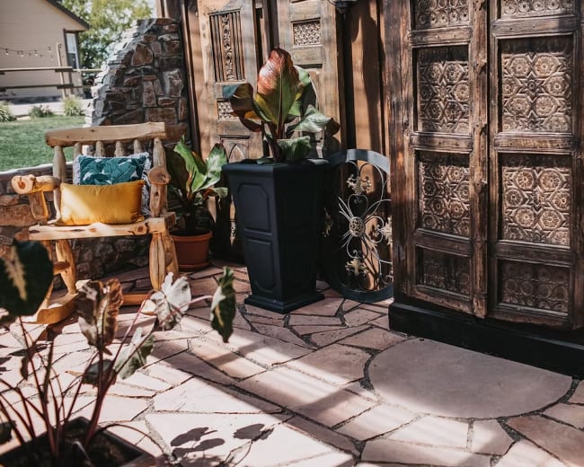 The image shows a cozy outdoor space featuring a stone-paved pathway, wooden furniture, potted plants, and intricately carved wooden doors.