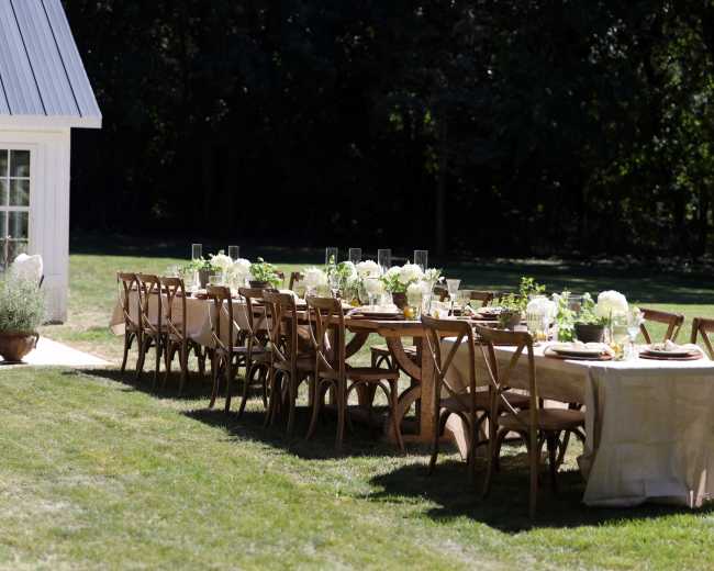 A long outdoor dining table is set with white flowers and tableware, surrounded by green grass and trees.