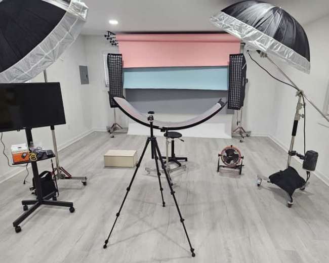 The image shows a photography studio setup with two softboxes, equipment stands, and a background featuring painted colors, along with a tripod and a television monitor.
