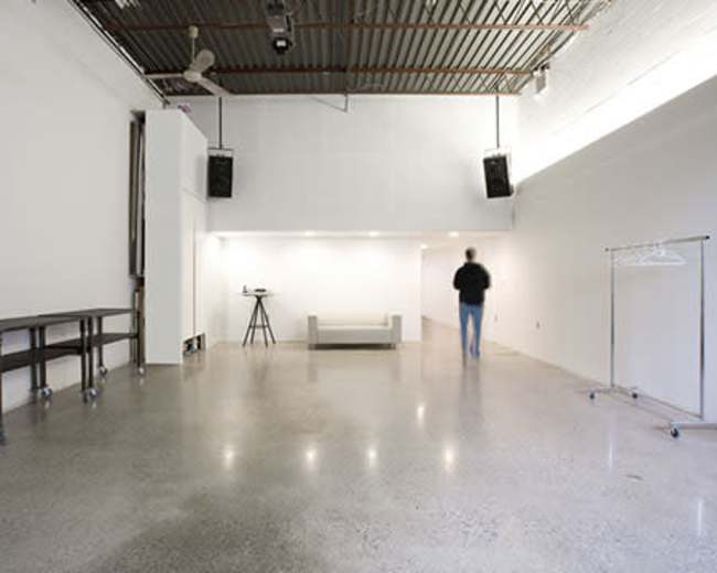 The image shows a spacious, minimalist room with polished concrete flooring, a sofa in the center, a standing clothing rack, and a person walking towards the back of the room.