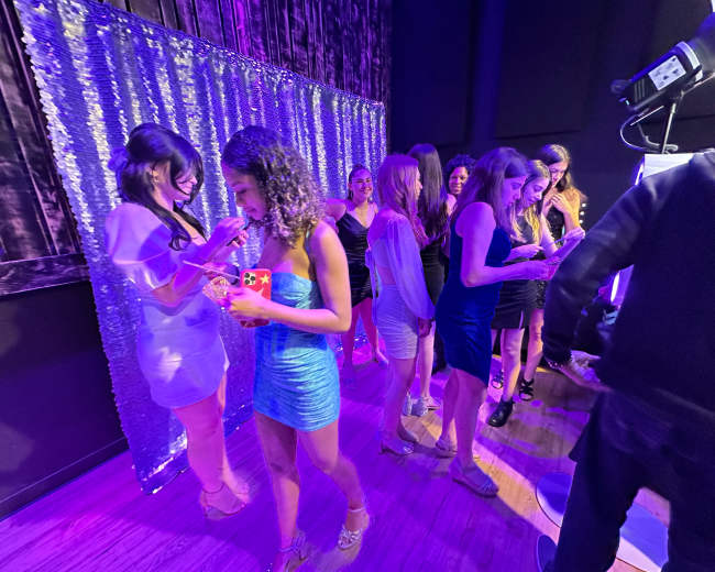 A group of women dressed in evening attire stands in front of a shimmering backdrop at a social event.