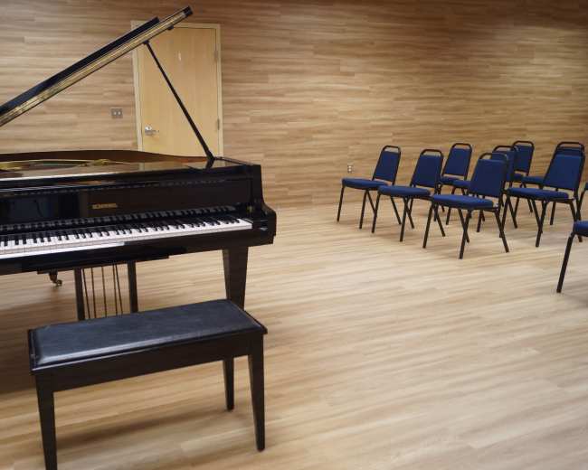 A black grand piano is positioned next to a black bench in a sparsely furnished room with wooden walls and empty blue chairs arranged in a semicircle.