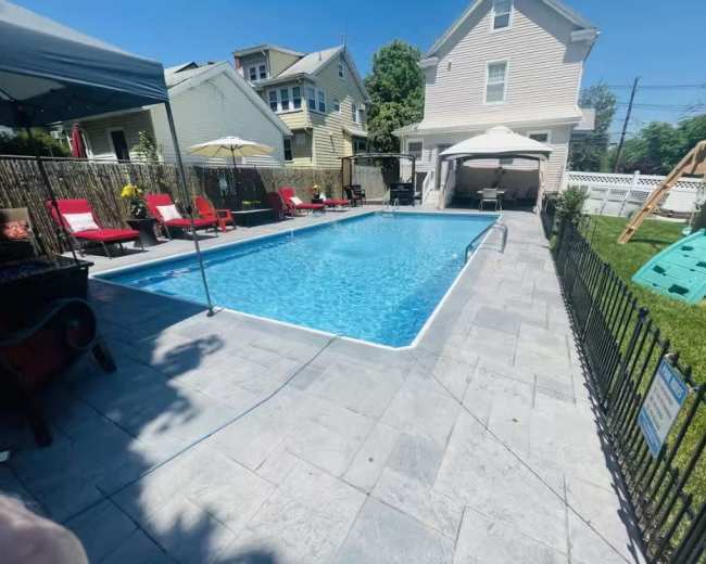The image shows a rectangular swimming pool surrounded by patio furniture and a grassy area, with a house and a playset in the background.