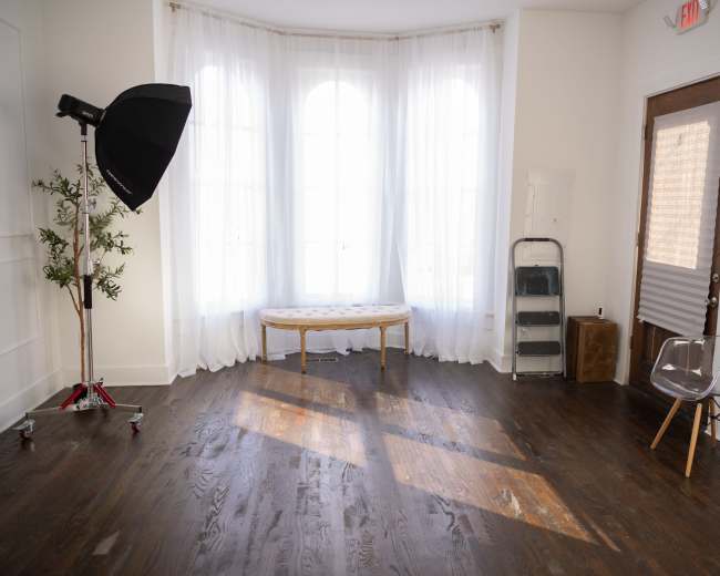 The image shows a well-lit room with large windows, a bench in the center, and a photography umbrella on a stand in one corner.