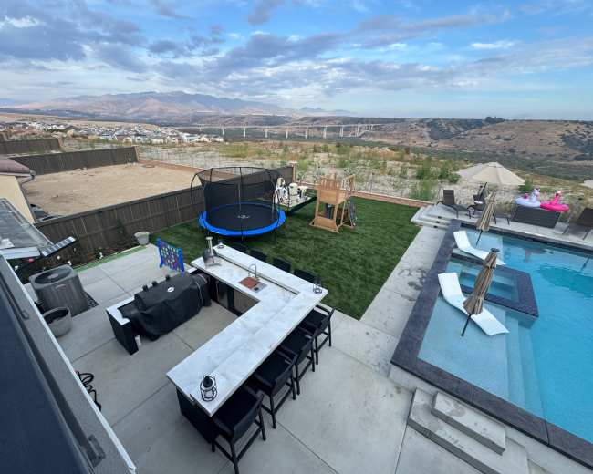 The image shows a backyard with a swimming pool, a patio with outdoor seating and a dining area, a trampoline, a play set, and a view of mountains in the distance.