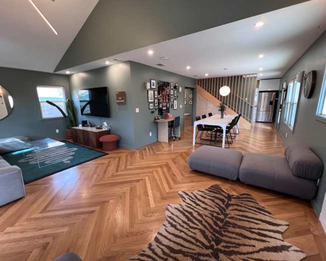 The image shows a modern living space featuring a comfortable seating area with a large rug, a dining table with chairs, a wall decorated with photos, and a staircase in the background.