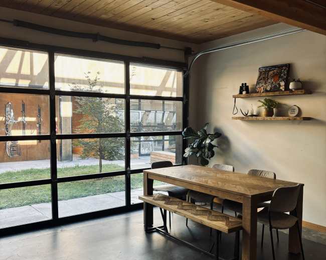The image shows a modern interior with a wooden dining table and chairs, large glass doors opening to an outdoor area, and a shelf mounted on the wall.