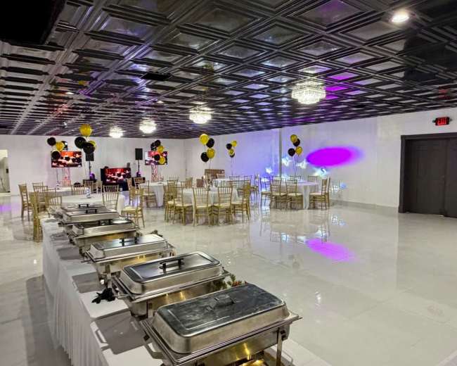 The image shows a spacious event hall set up for a celebration, featuring tables with white tablecloths, gold chairs, decorative balloons, and a buffet area with metal food warmers.