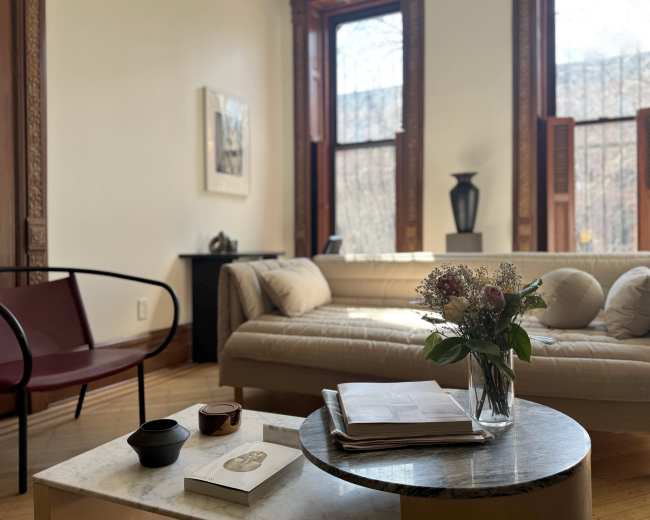 The image shows a well-lit living room with a beige sofa, a round marble coffee table adorned with magazines and a vase of flowers, and large windows with wooden frames.