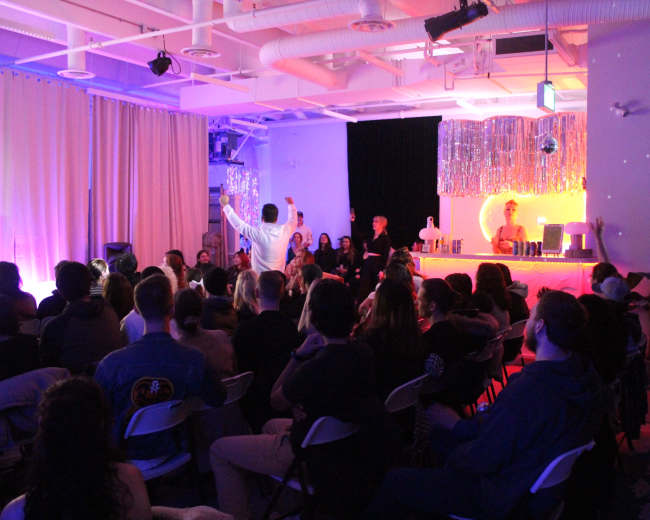 A crowd gathers in a brightly lit room, with a bar and a stage area visible in the background.