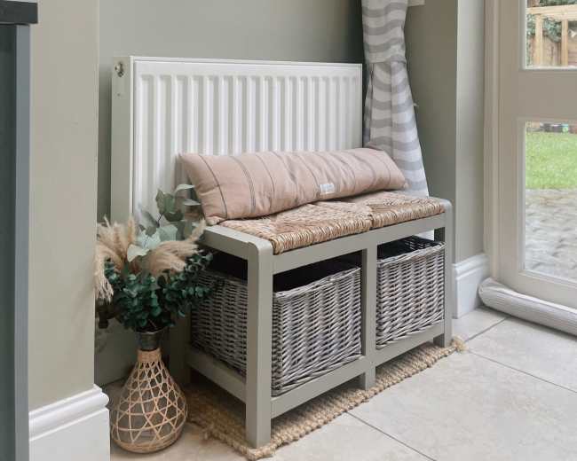 The image shows an entryway with a bench adorned with a cushion, two wicker baskets underneath, a wooden shelf above with a framed picture and a plant, and a window leading to a garden.
