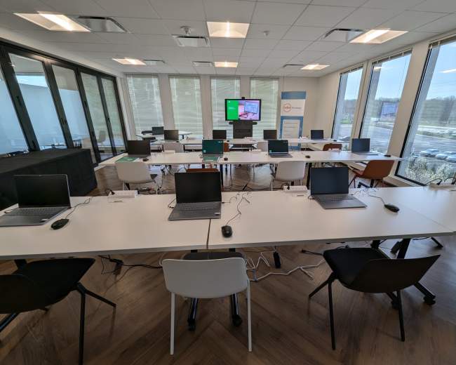 A modern classroom is set up with multiple tables, each featuring a laptop, mouse, and empty chairs, with large windows and a screen displaying information in the background.