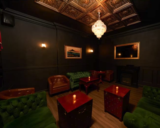 The image shows a darkly decorated lounge with green velvet seating, wooden tables, and a chandelier overhead.