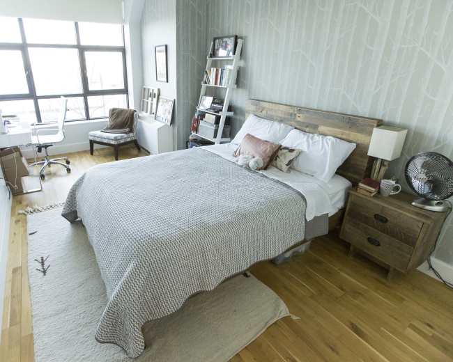 A neatly organized bedroom features a wooden bed with a light-colored blanket, a chair, a small desk, and large windows allowing natural light to enter.