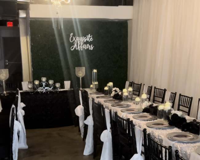 A long dining table adorned with white tablecloths and black accents is set up in a banquet space featuring greenery on the wall and a chandelier overhead.