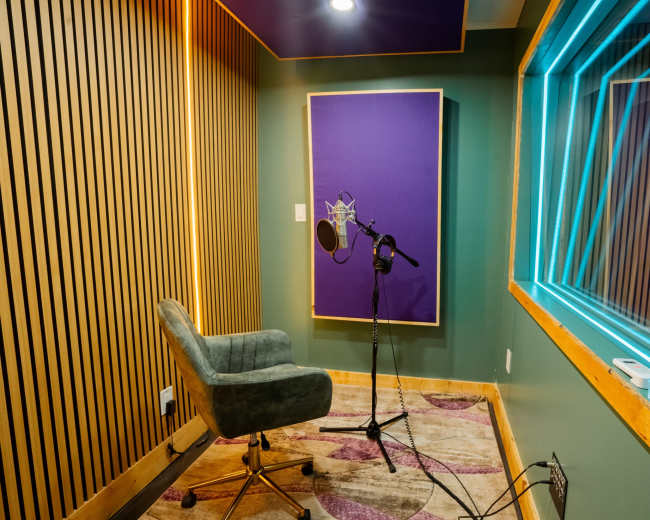 The image shows a sound recording booth with a microphone on a stand, a padded chair, and a glass window displaying a lighted geometric pattern.