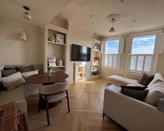 The image shows a cozy living room with a round dining table, a sofa, a television mounted on the wall, and large windows letting in natural light.