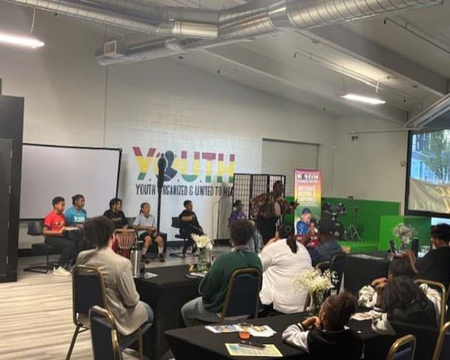 A group of young performers engages in a presentation on stage in front of an audience seated at tables in a community center.