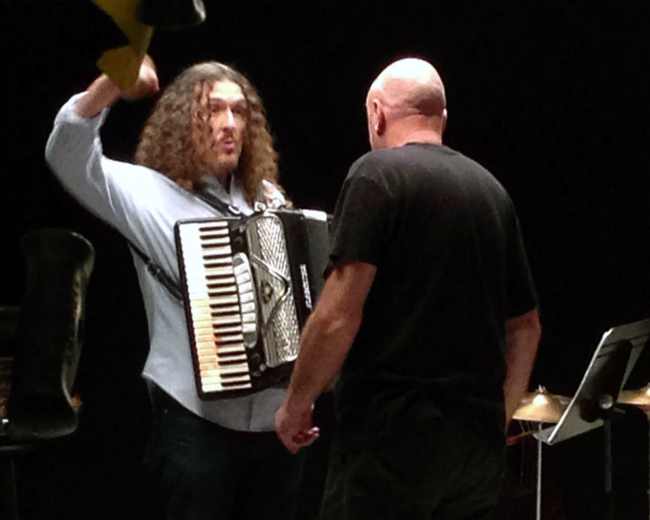 A man with curly hair plays an accordion while facing another man in a black shirt on a stage.