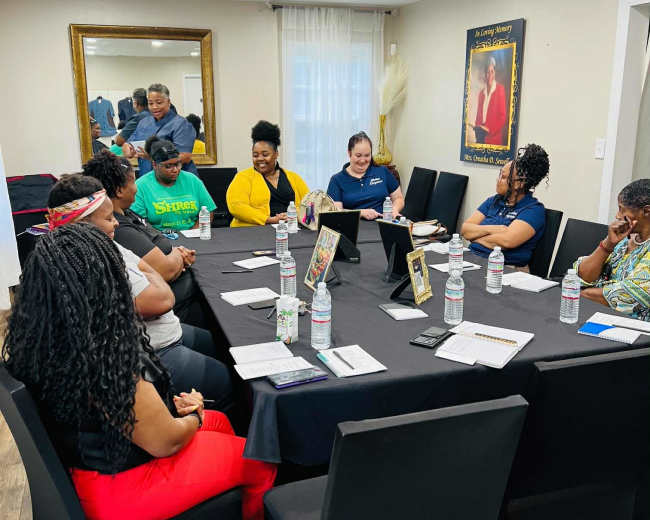 A group of eight individuals is seated around a rectangular table in a meeting room, engaged in discussion, with notes and water bottles in front of them.
