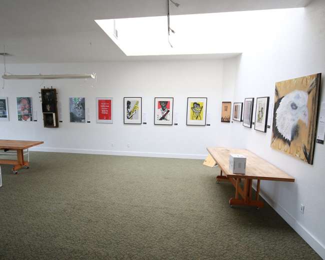 The image shows an art gallery with a person seated at a table while various framed artworks hang on the white walls.