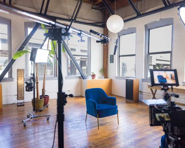 The image features a well-lit filming studio with a blue chair set up in front of a camera and lighting equipment, surrounded by large windows.