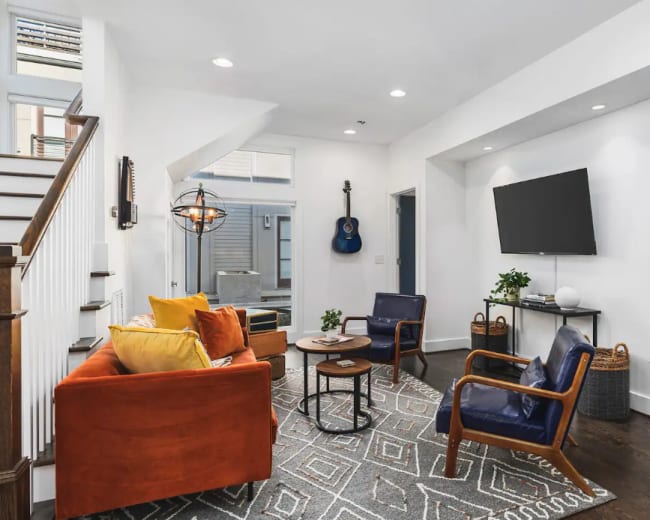 The living room features a mix of orange and blue armchairs, a round coffee table, and a mounted television against a white wall, with a guitar hanging on the adjacent wall.
