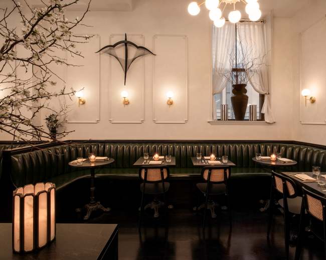 The image shows a dimly lit restaurant interior featuring green leather banquettes, wooden tables, and decorative wall accents, including a large vase and an umbrella display.