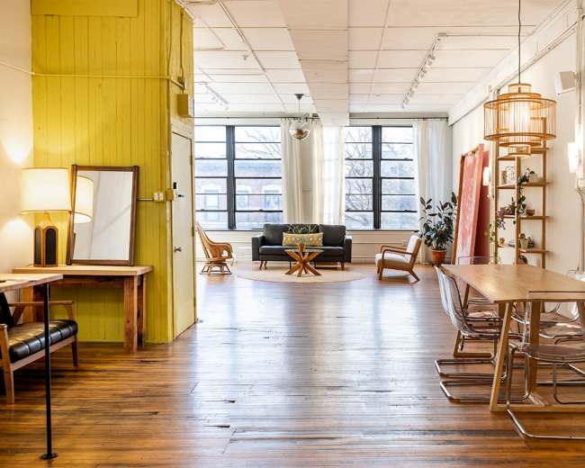 The image shows a spacious open-plan room with wooden flooring, featuring a seating area with a sofa and decorative plants, a dining table, and large windows allowing natural light to enter.