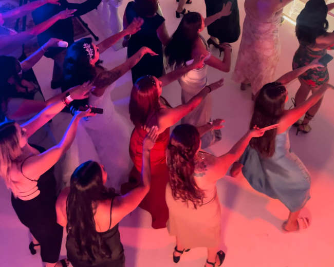 A large group of people is participating in a dance activity under colorful lights and decorative chandeliers.