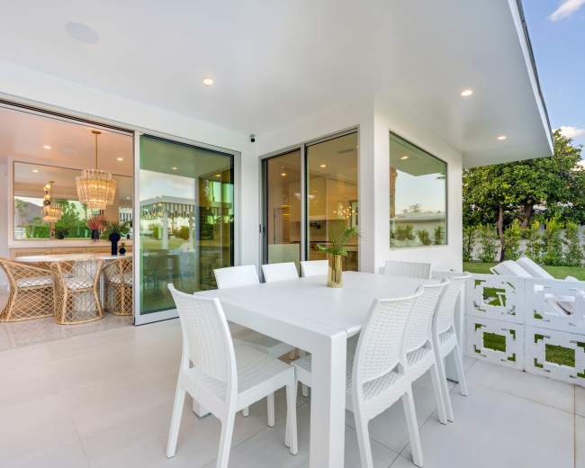 The image shows a modern patio with a white dining table and chairs, adjacent to an interior dining area featuring a chandelier and large windows.