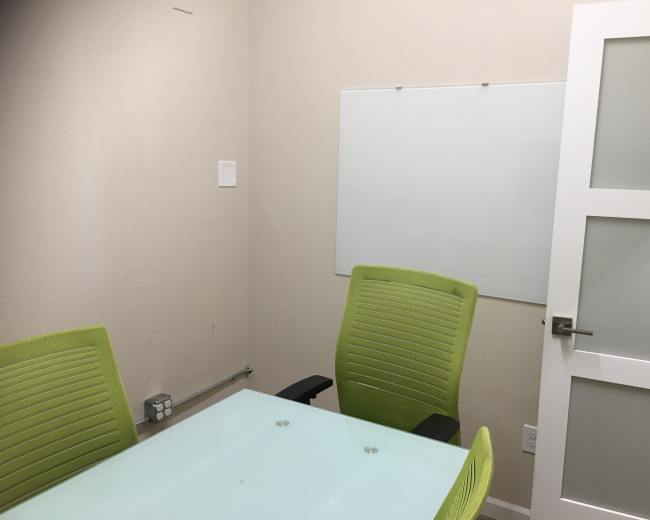 A small conference room with a glass table, two green chairs, and a whiteboard on the wall.