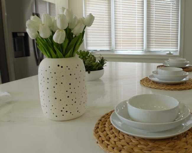 A dining area features a table set with white bowls on woven placemats, accompanied by a vase of tulips and a small plant in the background.