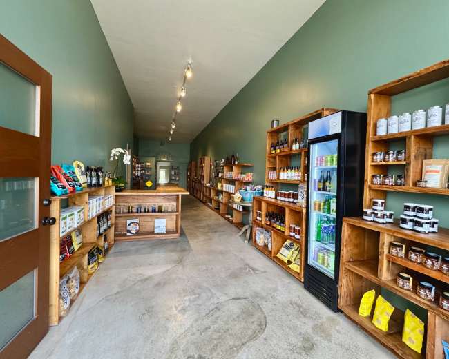 The image shows the interior of a small store featuring wooden shelves filled with various packaged goods and a refrigerator displaying beverages along one side.