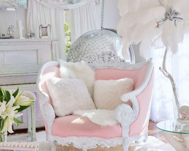 The image shows a bright interior featuring a pink upholstered sofa, white fur pillows, a glass table, and a large disco ball surrounded by crystal chandeliers.