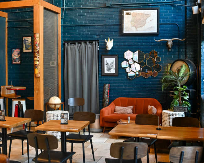 A cozy café features wooden tables and chairs, a bright orange couch, and a blue brick wall adorned with framed artwork and decorative elements.