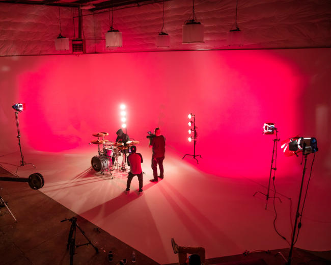 A band performs on a stage with a bright pink backdrop and various lighting equipment set up around them.
