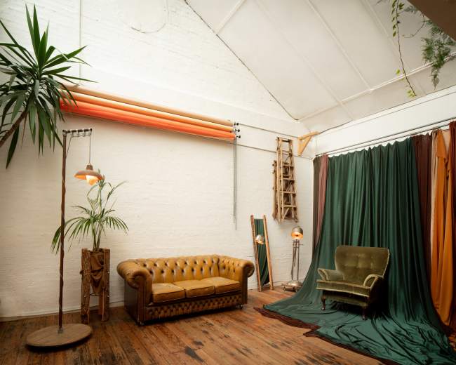 The image shows an interior space featuring a vintage brown leather couch, a green velvet armchair, a standing lamp, and draped fabric in various colors against a white brick wall.