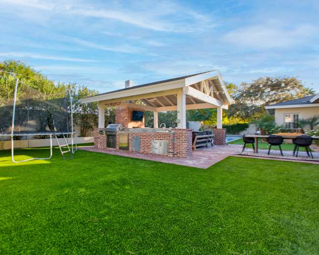 A backyard with a patio featuring a brick barbecue grill and seating area, surrounded by lush green grass and a trampoline.