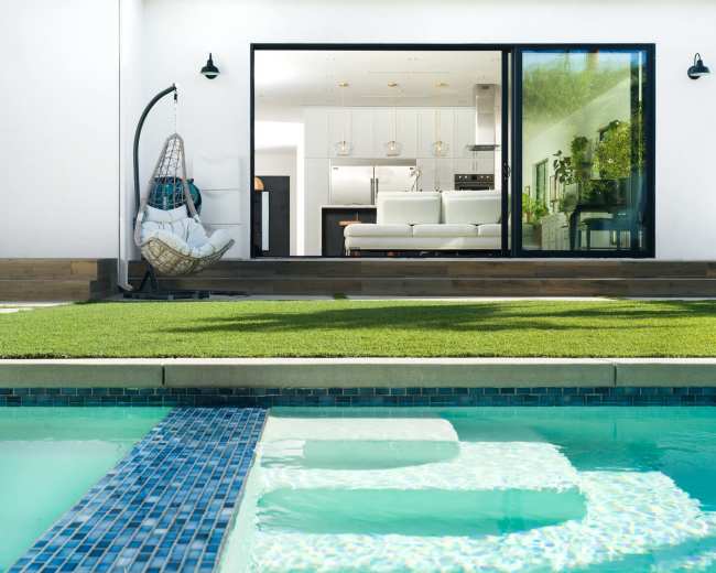 A glass sliding door opens from a modern living space to a backyard with a pool and a neatly manicured lawn.