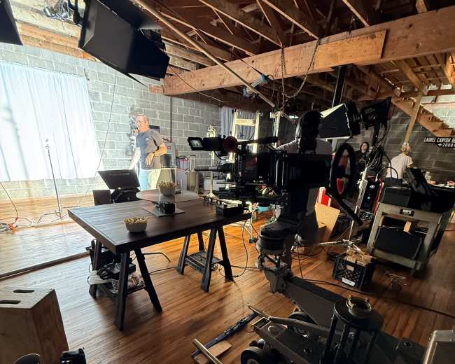 The image shows a film set with various lighting equipment, a camera on a dolly, and a crew member preparing near a table in a wooden loft space.