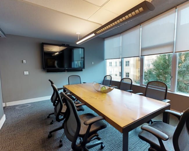 A conference room featuring a long wooden table surrounded by black office chairs and large windows illuminating the space.