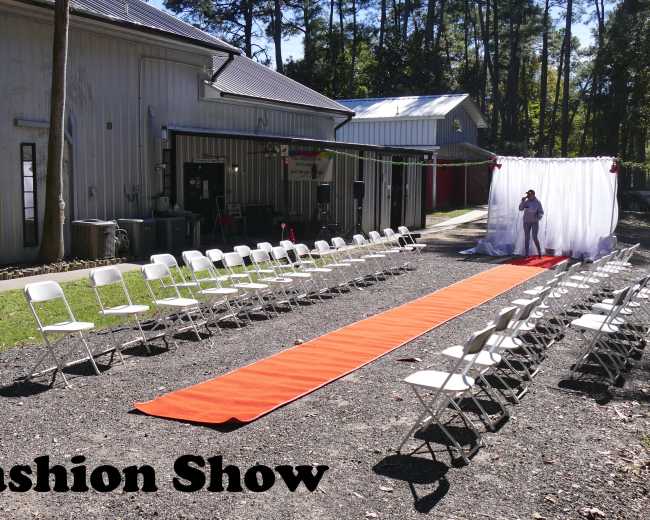A fashion show setup features a long orange runway surrounded by white chairs, with a backdrop of curtains at one end.