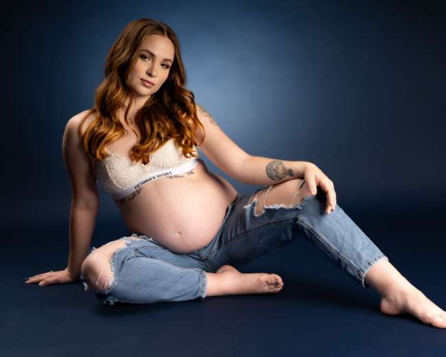 A pregnant individual poses seated on a blue background, wearing a lace bralette and distressed jeans.