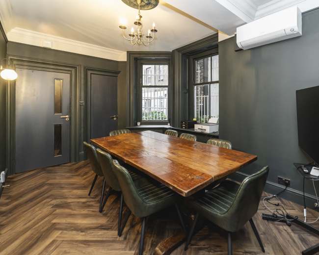 The image shows a dining area with a large wooden table surrounded by green chairs, and a flat-screen TV mounted on a wall in a room with dark green walls and large windows.