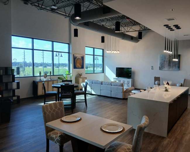 The image shows a modern open-concept living space featuring a kitchen island, dining area, and a seating area with large windows providing natural light.
