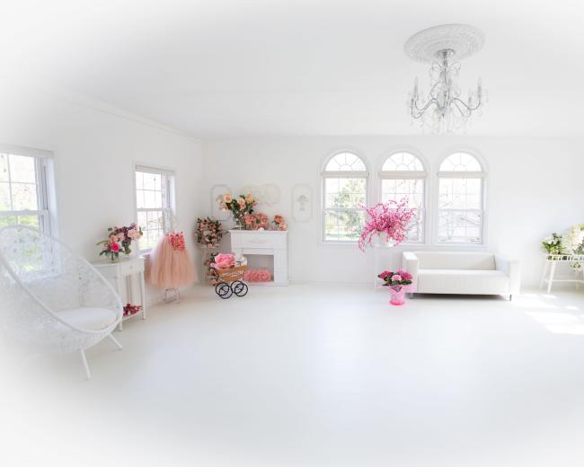 The image depicts a bright, airy room with white walls and elegant decor, featuring large windows, a chandelier, and various floral arrangements.