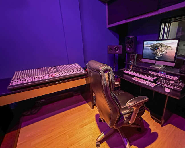 A music studio with a mixing console, computer, and sound equipment, illuminated by purple lighting.
